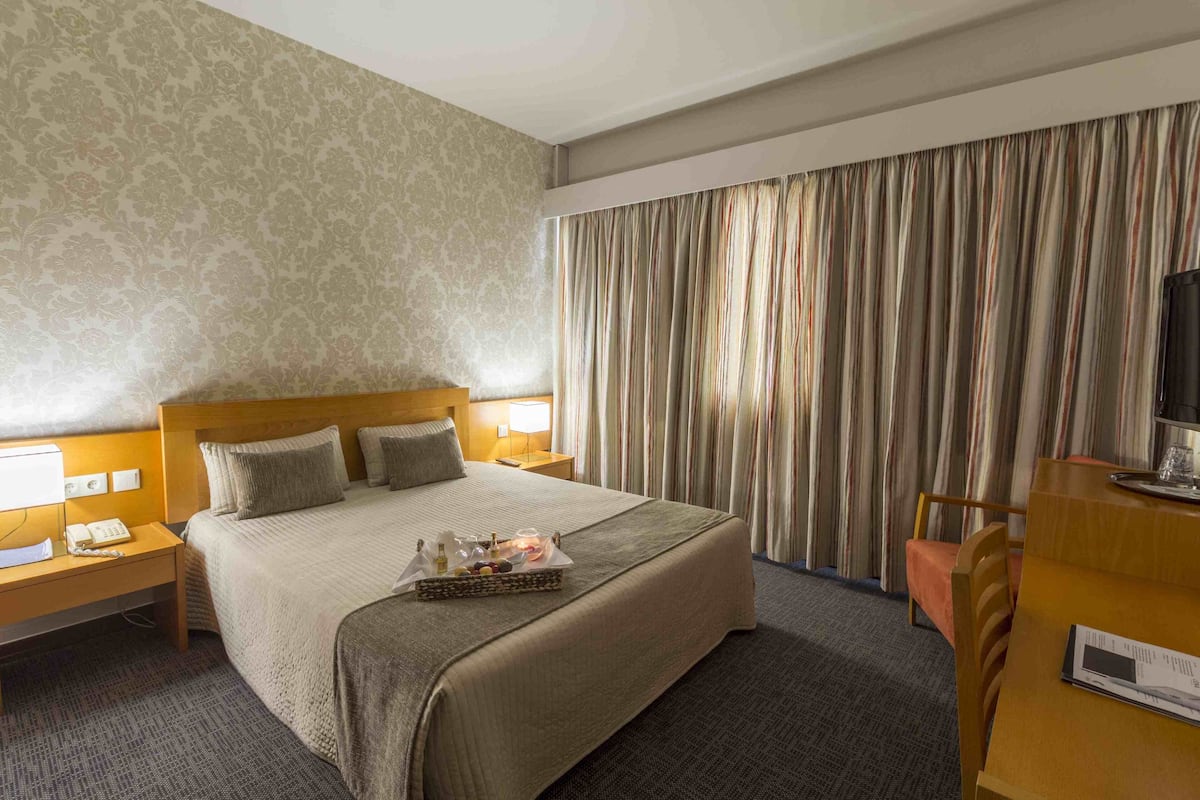 A hotel room with a large bed, two bedside tables with lamps, a wooden desk, and a large window with curtains.