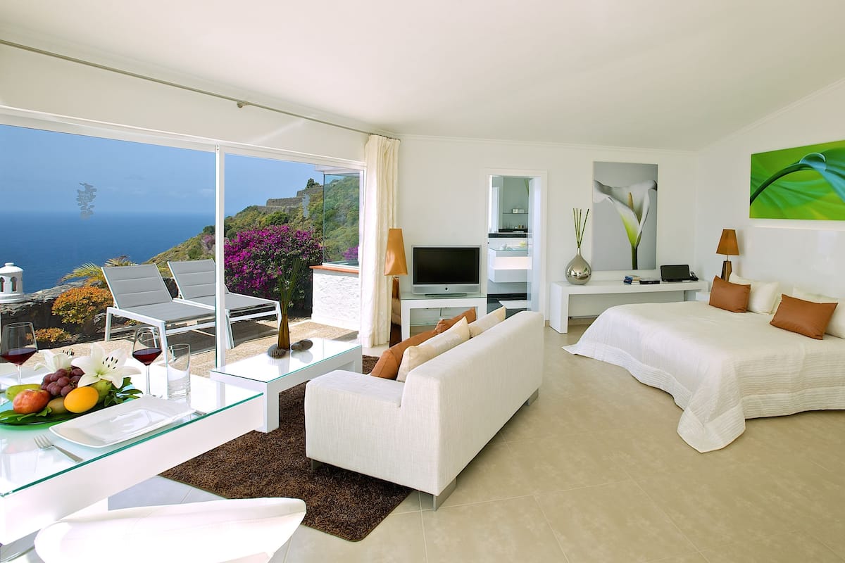 Habitación de hotel moderna con balcón, cama, sofá, televisor y zona de comedor con una mesa para dos.