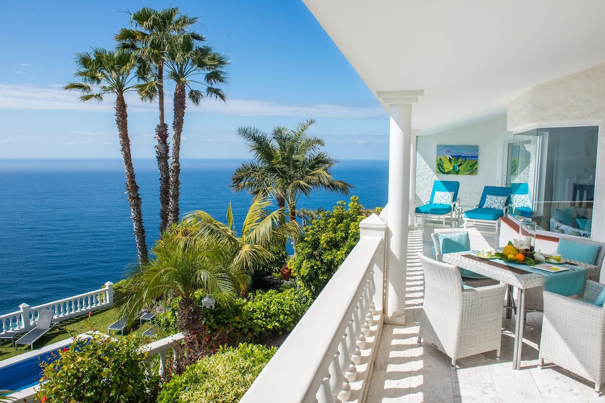 Un balcón con mobiliario de mimbre blanco, piscina y vista al océano.