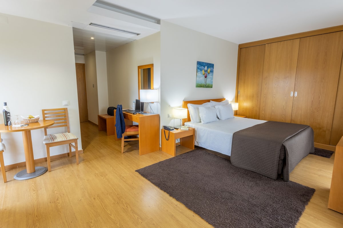 Habitación de hotel con cama, escritorio con silla, mesita de noche con lámpara y armario.