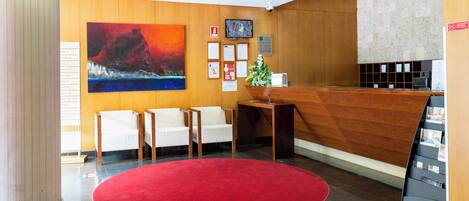 Un área de recepción con un mostrador de madera, alfombra roja, sillas blancas y un cuadro grande en la pared.