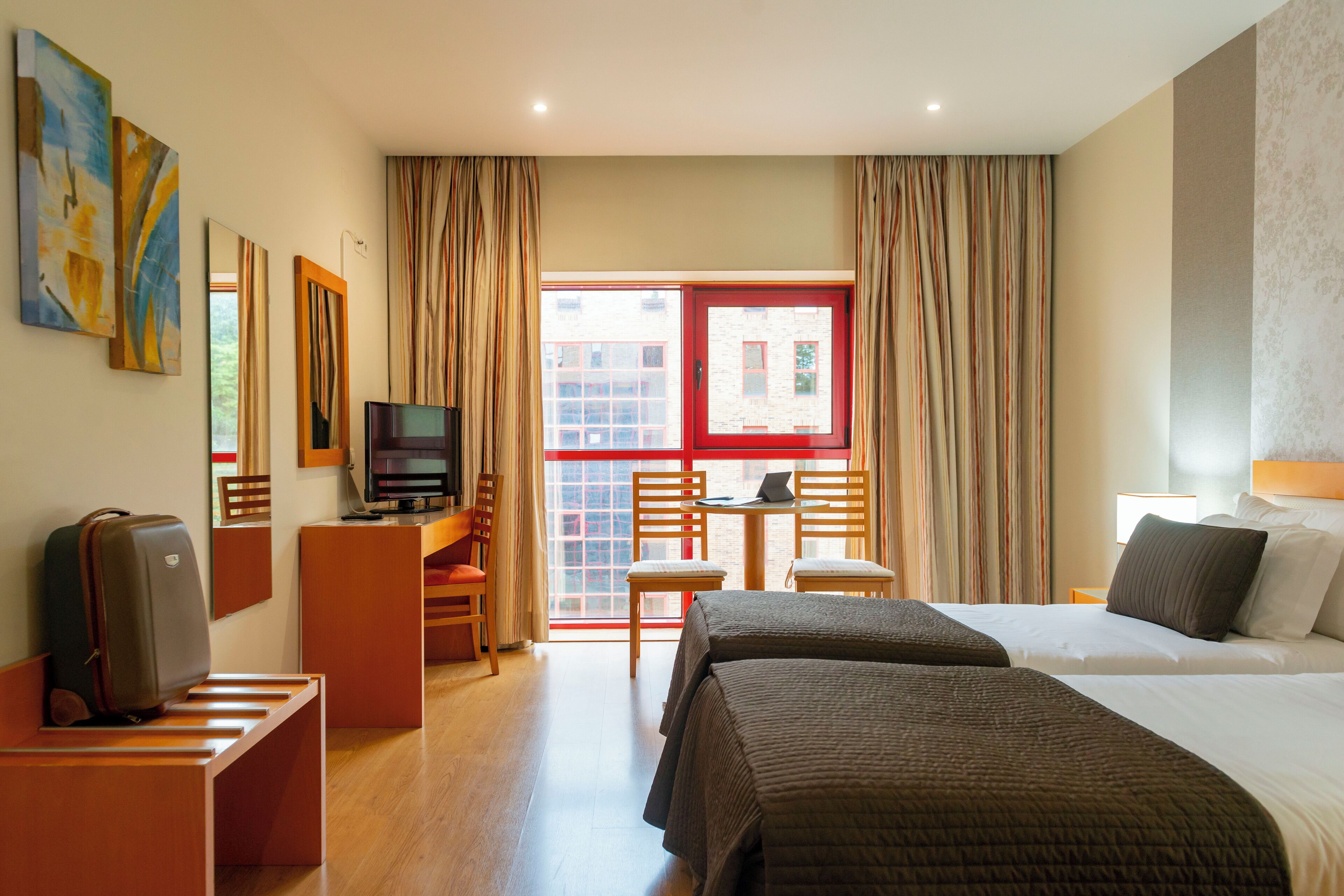 A hotel room with two beds, a desk with a chair, a television, and a suitcase on a bench.