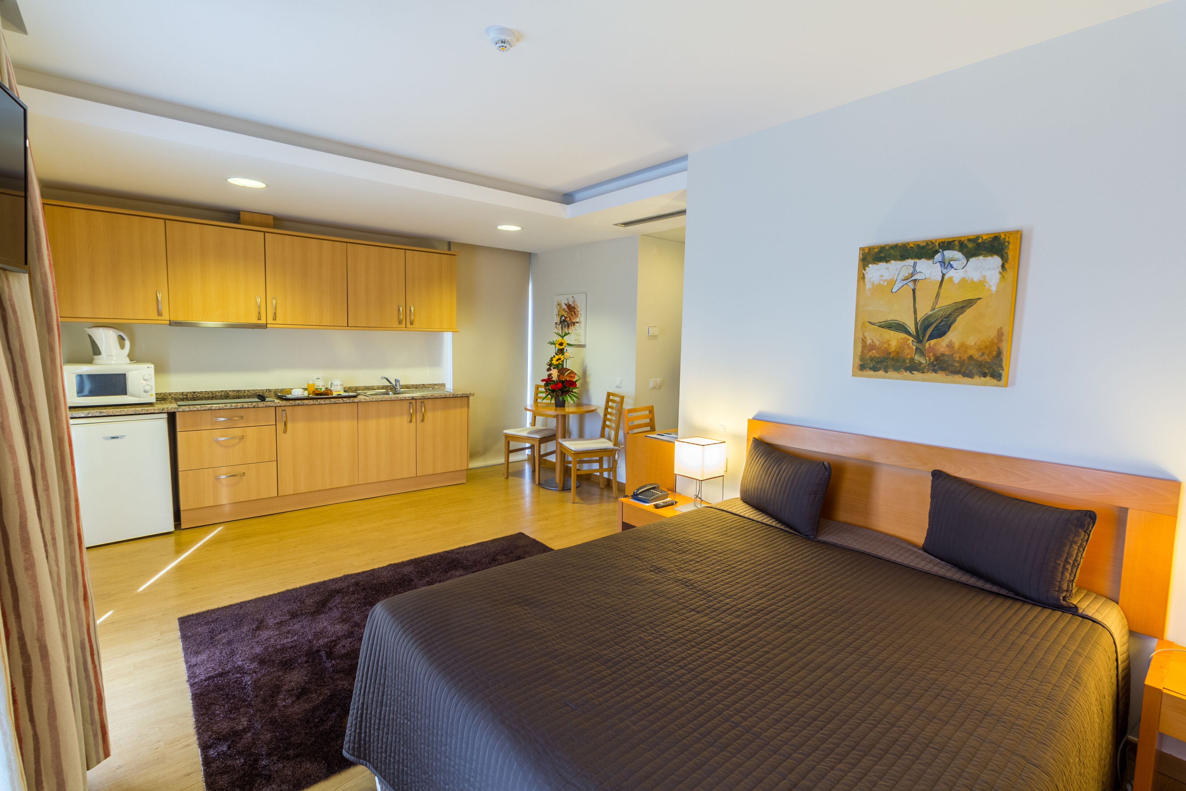 A modern hotel room with a large bed, a wooden headboard, a small desk, and a kitchenette in the background.
