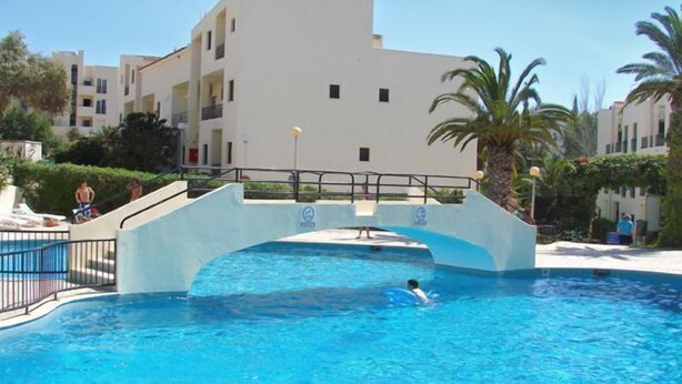 A swimming pool with a bridge, surrounded by buildings and palm trees. - Clube Alvorferias (Portimão)