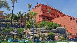 A hotel with a red facade, multiple balconies, and a pool area with lounge chairs. - Apartamentos Manilva Green (Manilva)