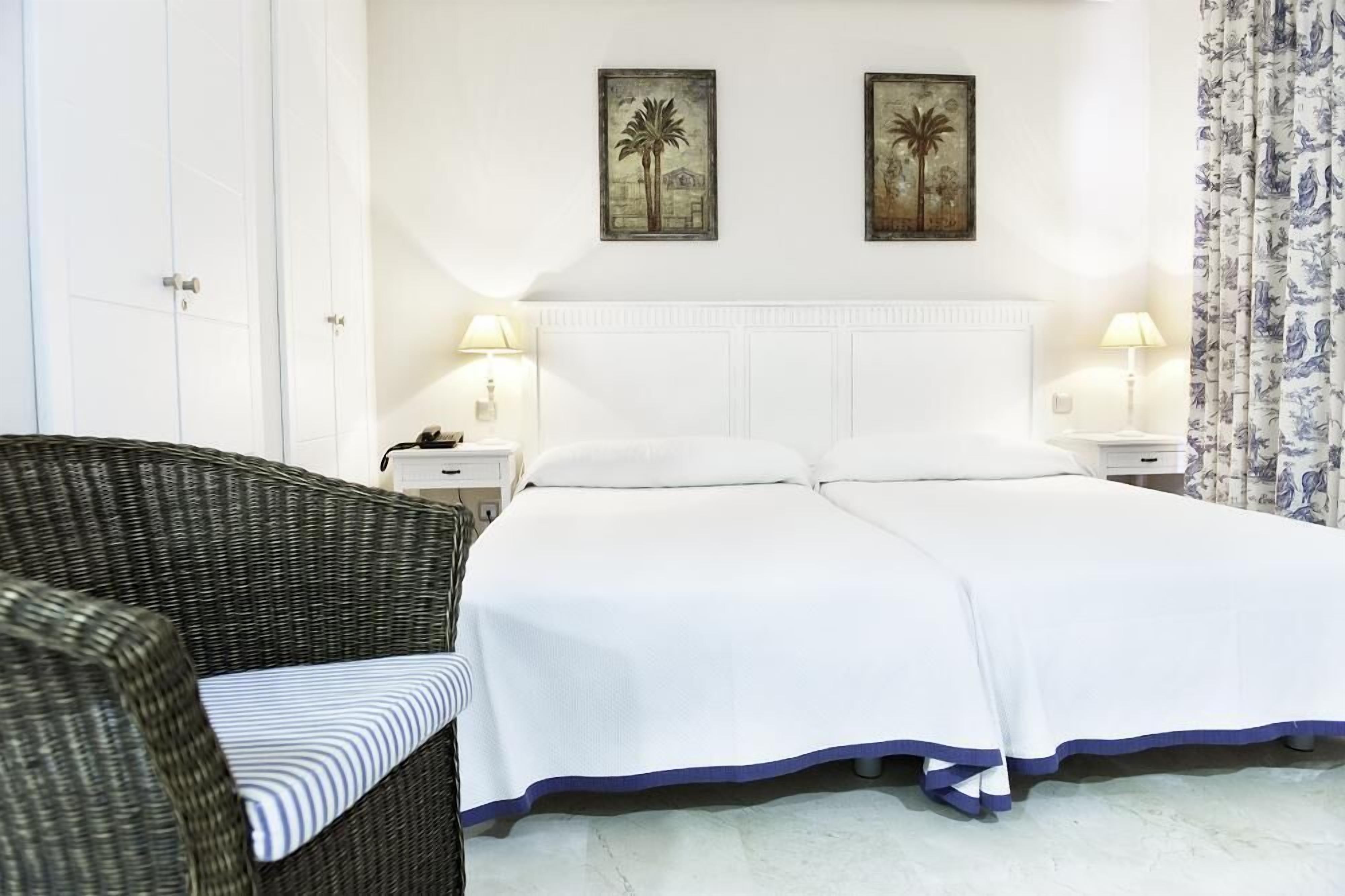 A bedroom with a bed, two nightstands, a wicker chair, and two wall-mounted paintings.