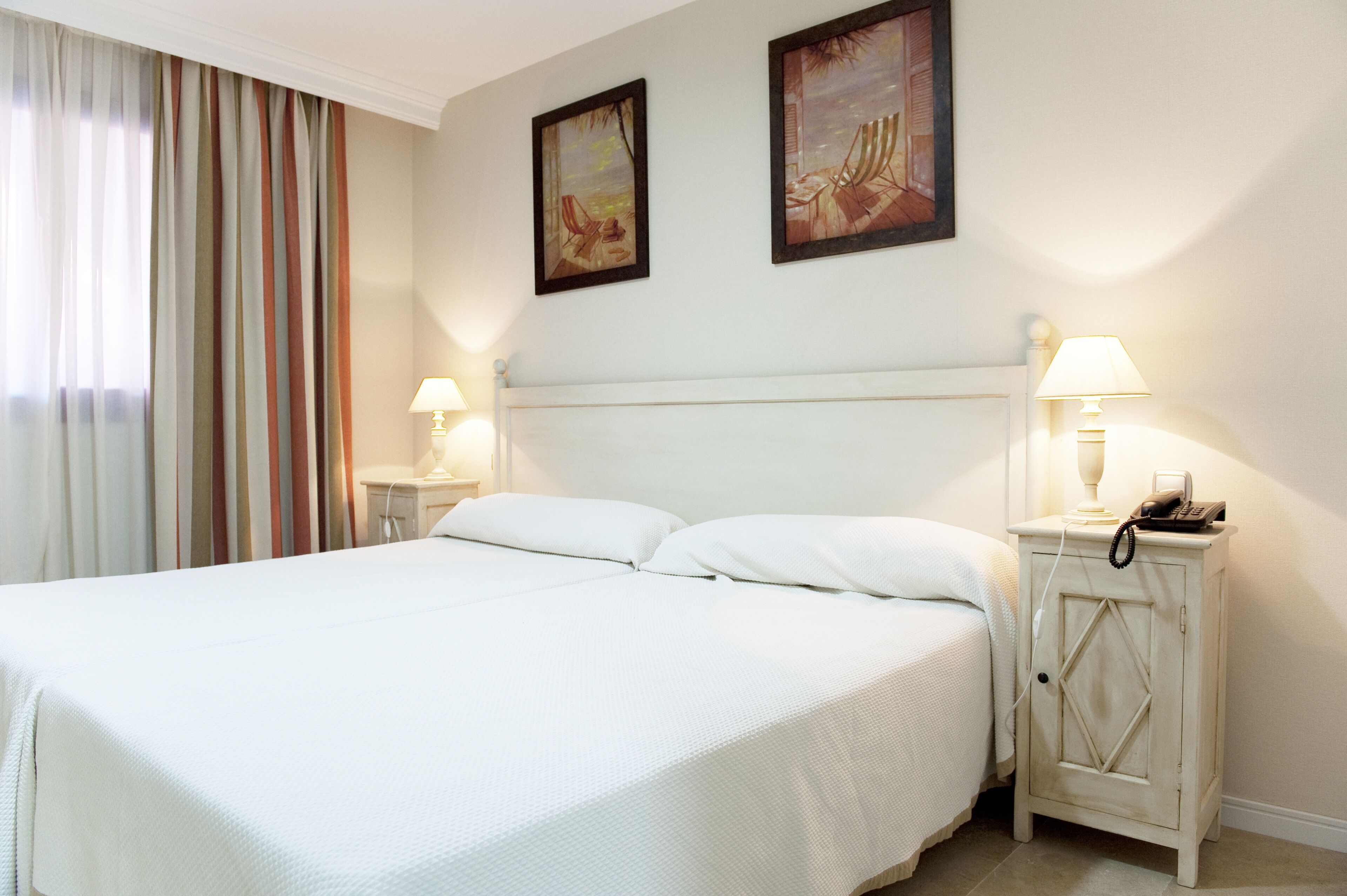 Una habitación de hotel con una cama grande, dos mesitas de noche con lámparas y dos cuadros en la pared.