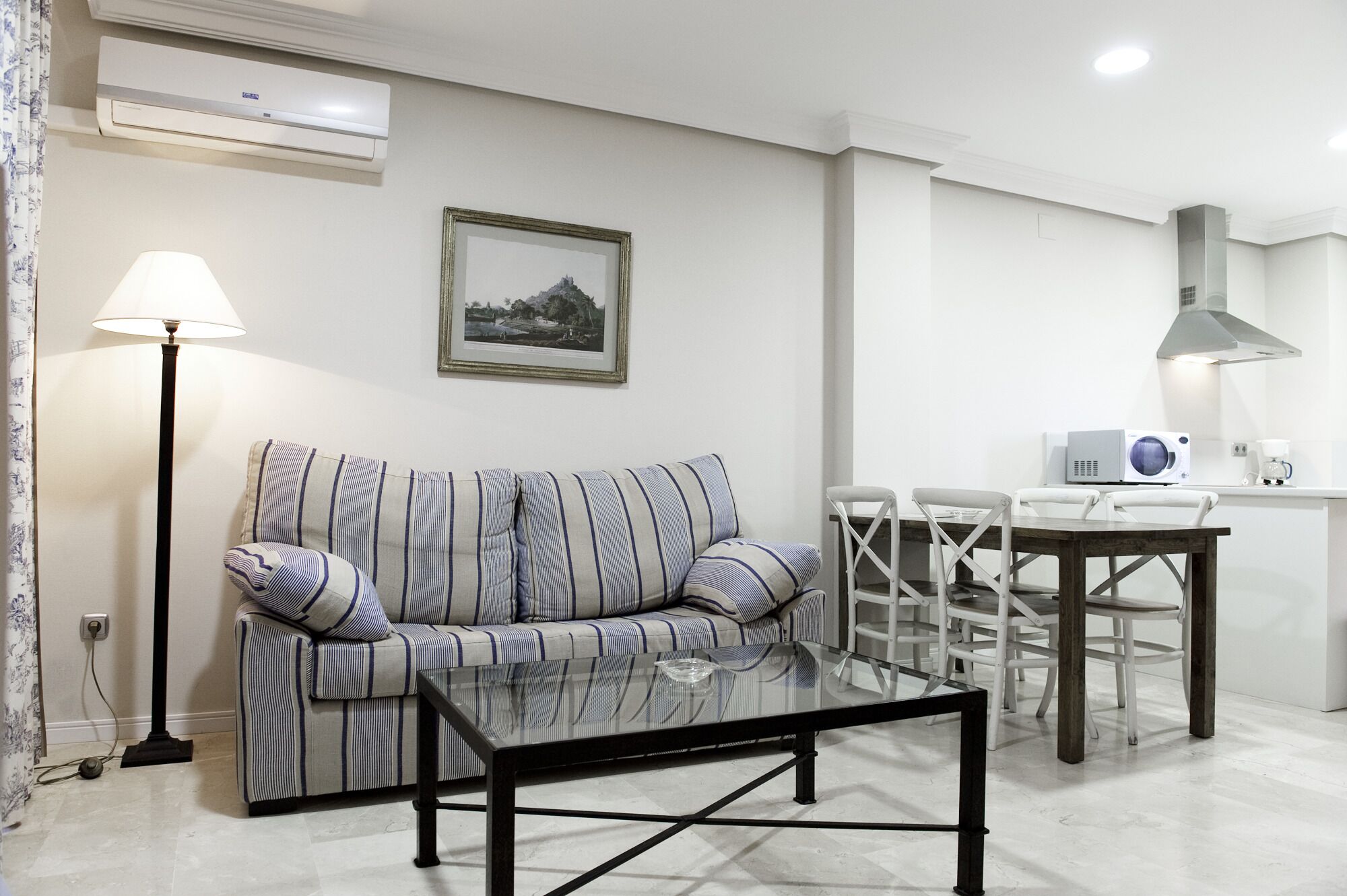 Un salon avec un canapé à rayures, une table basse en verre, une table à manger avec des chaises, un four à micro-ondes et un tableau accroché au mur.