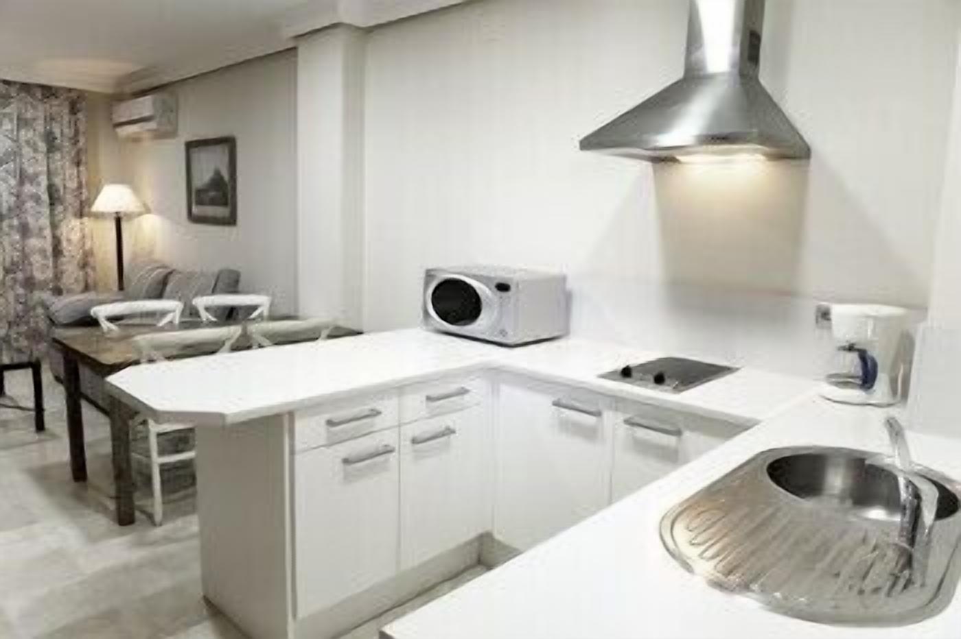Een compacte keuken met witte kasten, een ingebouwde magnetron, een spoelbak en een klein eethoekje met een tafel en stoelen.