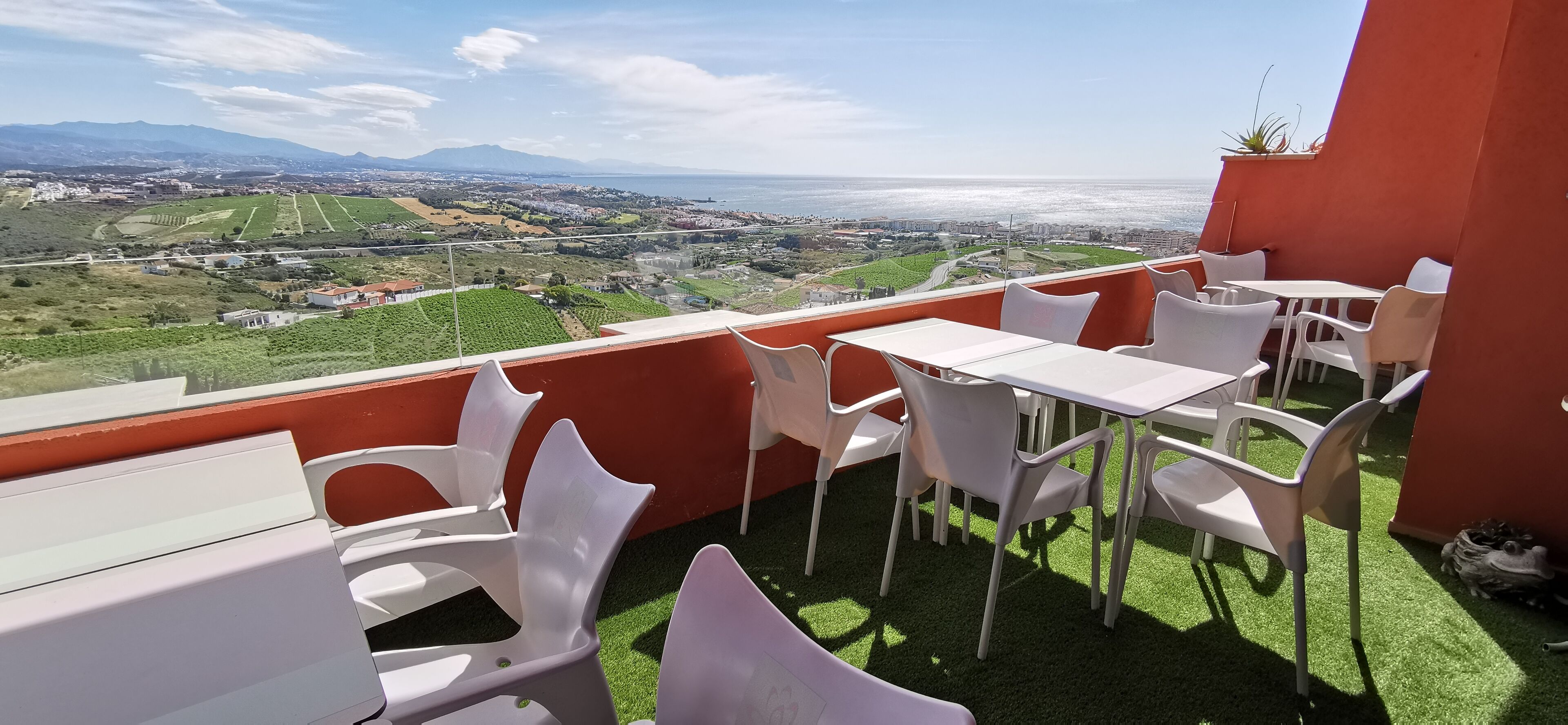 A terrace with white plastic chairs and tables, overlooking a coastal landscape with fields and buildings.