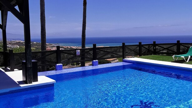 A clear blue pool with a view of the ocean, surrounded by palm trees and a wooden deck. - Apartamentos Manilva Green (Manilva)