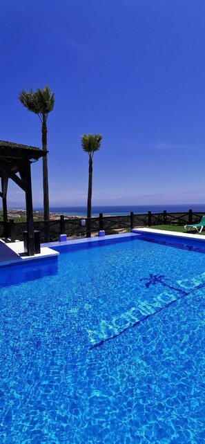 Piscina azul y cristalina con vista al océano, rodeada de palmeras y una terraza de madera.