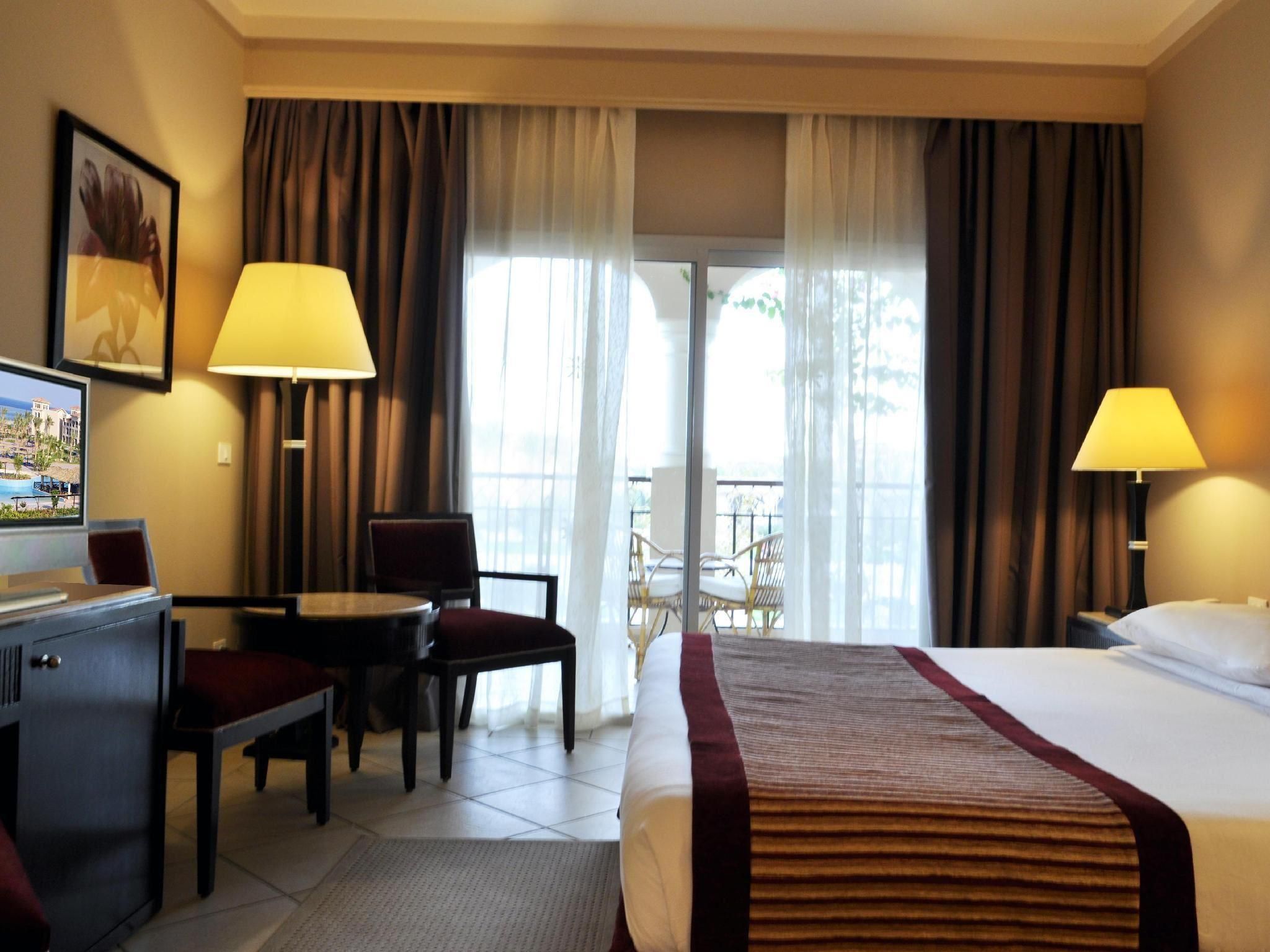 A hotel room with a bed, two bedside lamps, a desk with a chair, a television, and a balcony with chairs.