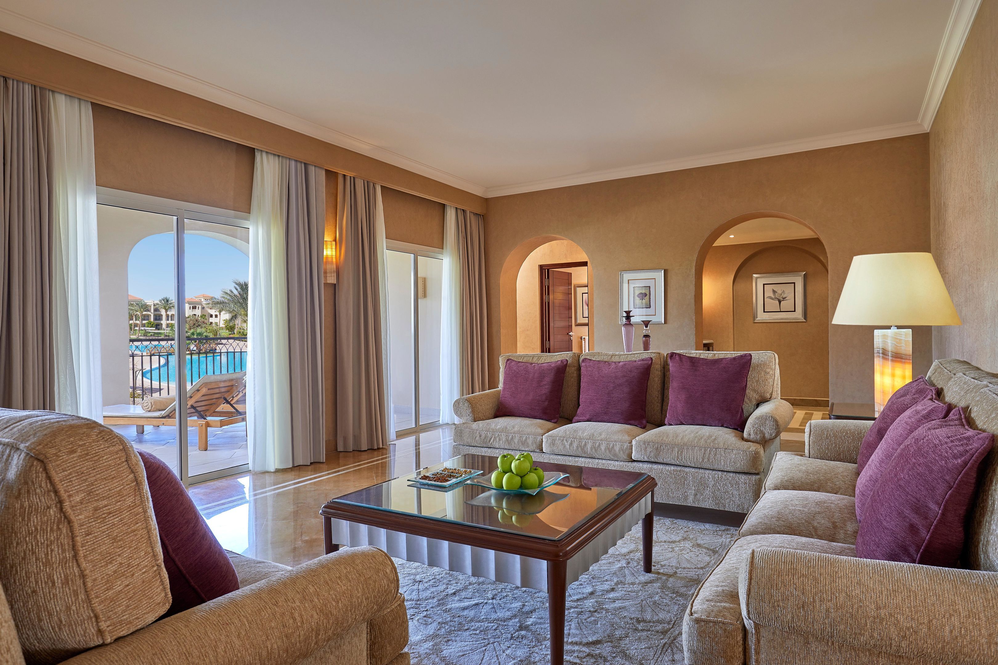 A hotel room with a sofa, armchairs, a glass coffee table, and a view of a pool.