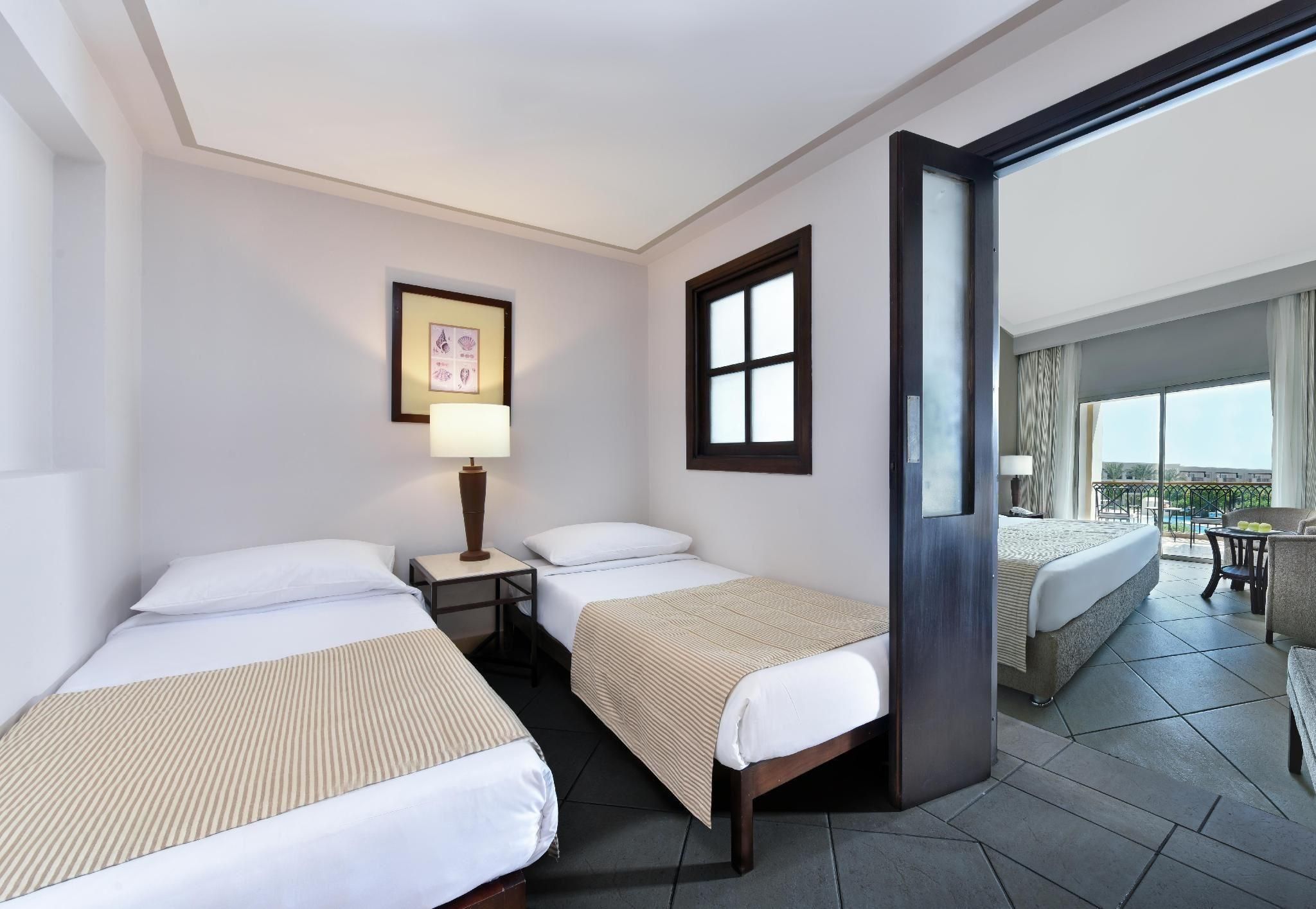 Habitación de hotel con dos camas individuales, una mesita de noche con lámpara, un cuadro en la pared y un balcón con mesa y sillas.
