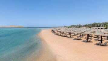 Une plage bordée de rangées de chaises longues et de parasols, une mer d’un bleu limpide et une jetée qui s’étend au loin.