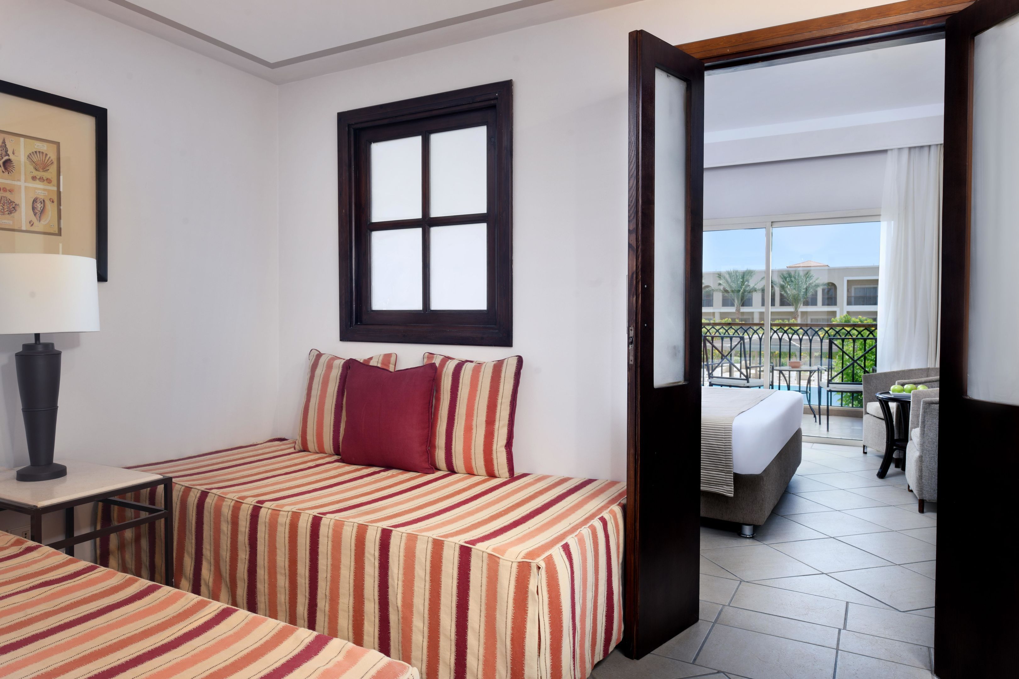 A hotel room with a bed, a small table, a lamp, a window, and a balcony with outdoor furniture.