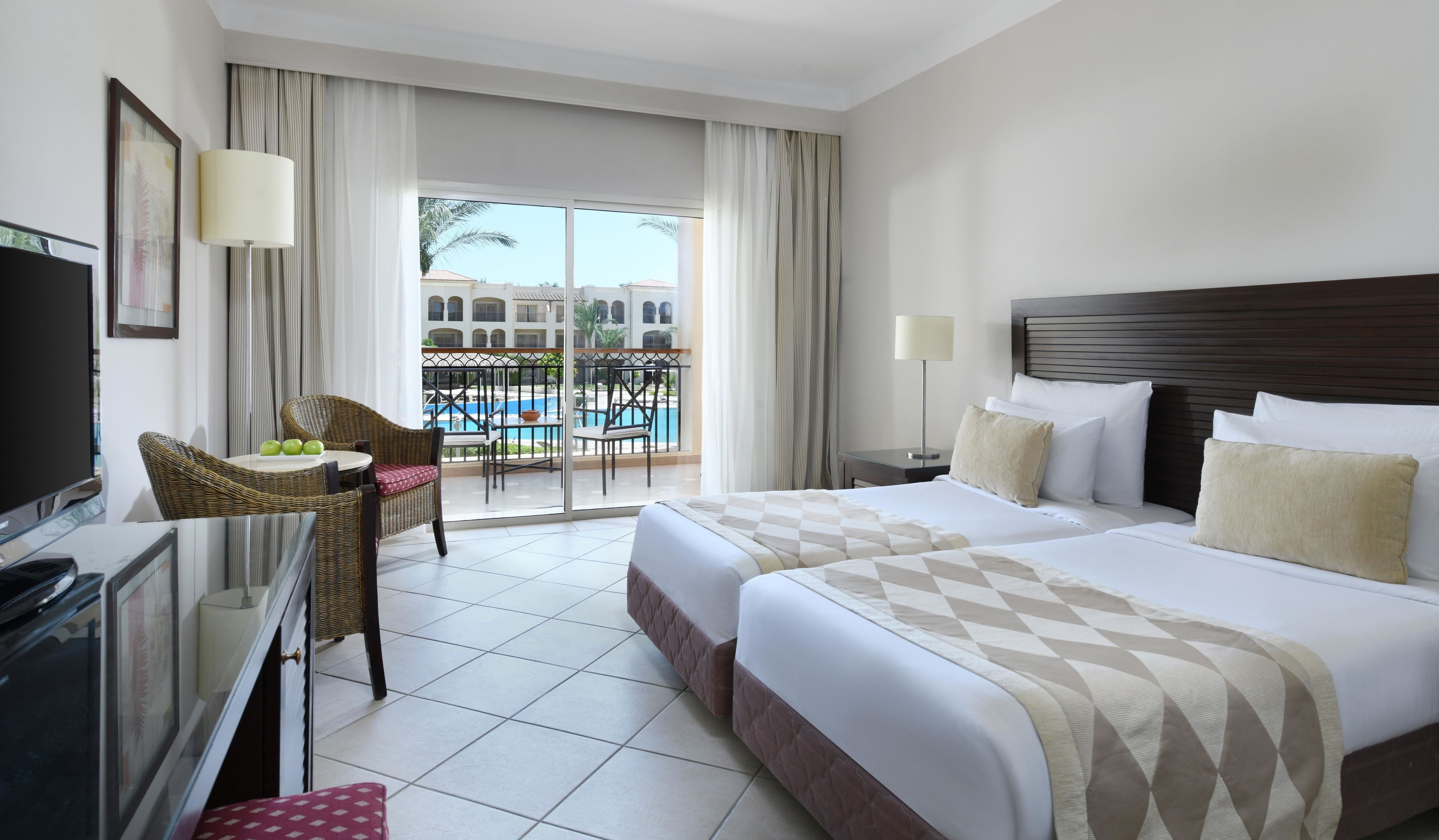 Habitación de hotel con cama, televisión, escritorio, silla, balcón con vista a la piscina y ventana con cortinas.