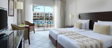 A hotel room with a bed, a TV, a desk, a chair, a balcony with a pool view, and a window with curtains.