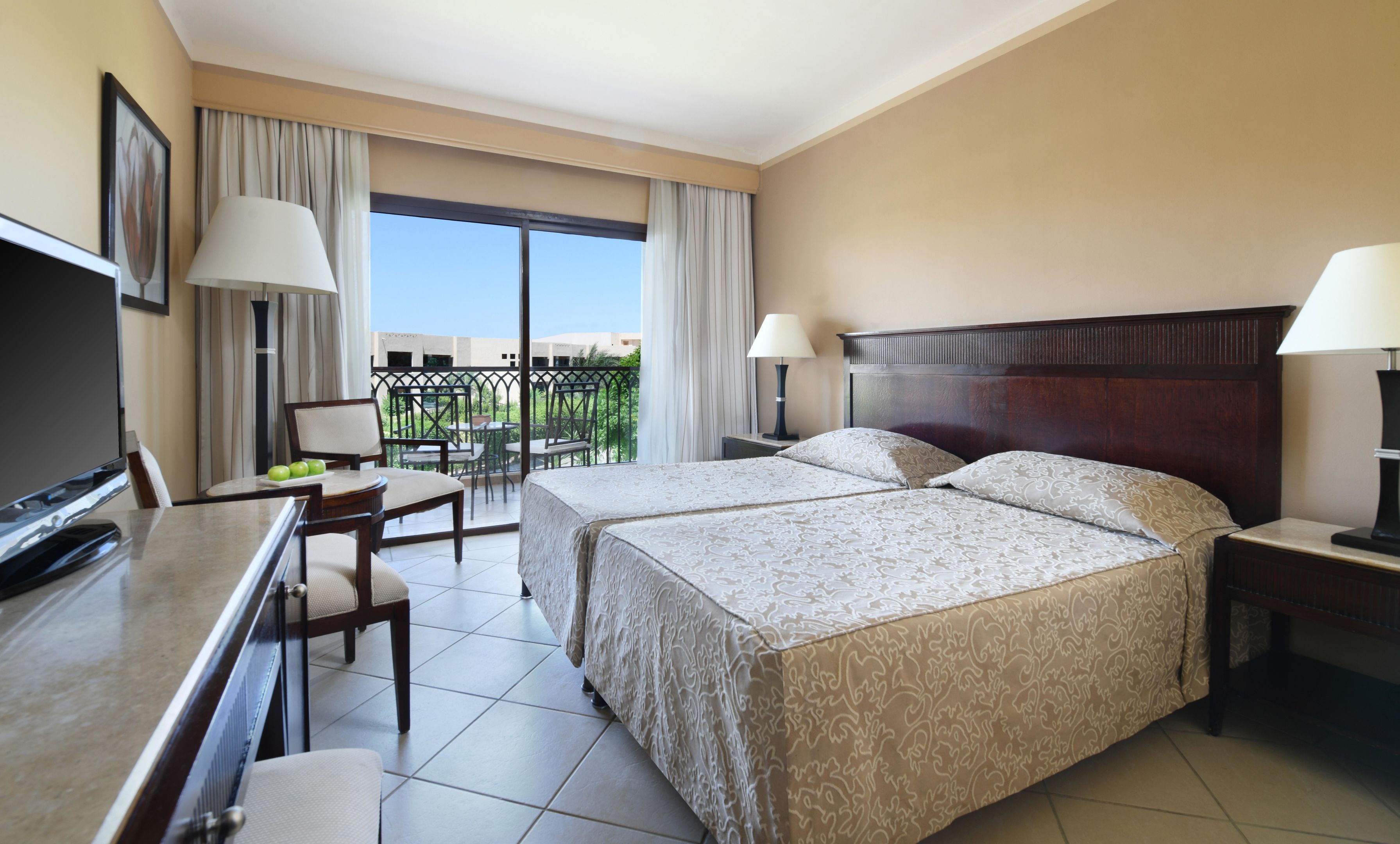 Habitación de hotel con dos camas, un escritorio con televisor, un balcón con vistas y una zona de comedor con sillas.