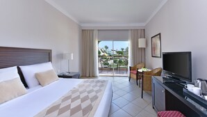 A hotel room with a bed, a TV, a desk, and a balcony with a view of a water park. - JAZ Mirabel Resort – All Inclusive (Sharm El Sheikh)