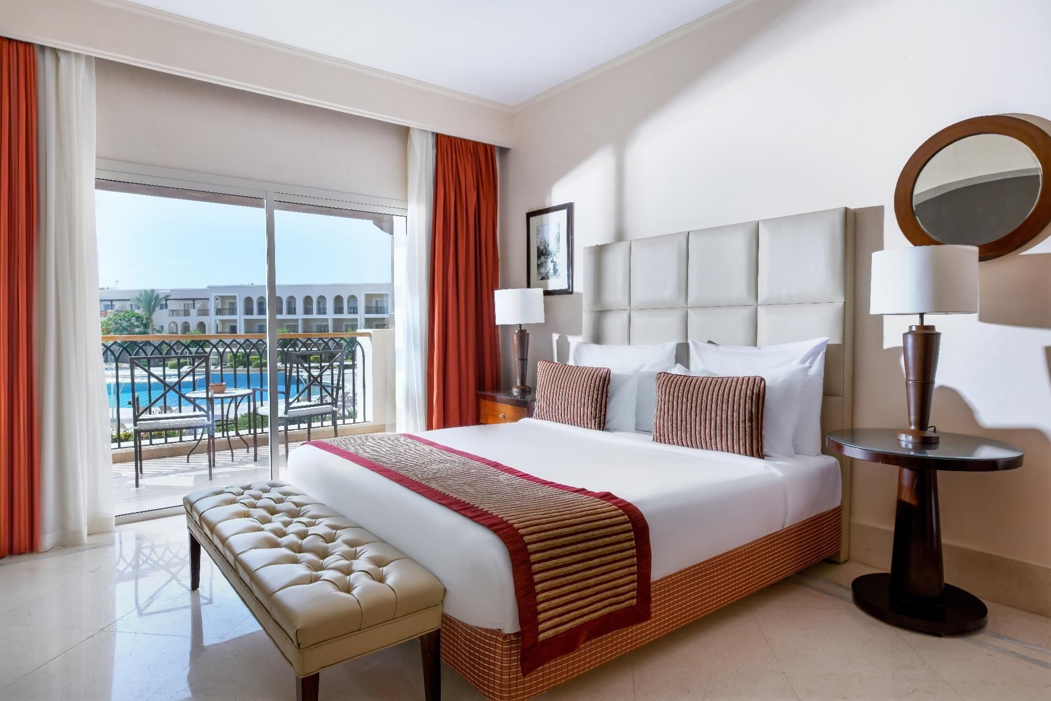 Une chambre d’hôtel avec un grand lit, un banc, une table ronde et une vue sur une piscine et des immeubles.