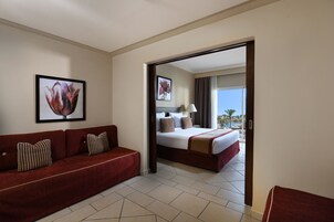 A hotel room with a red sofa, a bed with white bedding, a framed floral artwork on the wall, and a view of a pool and palm trees through an open door.