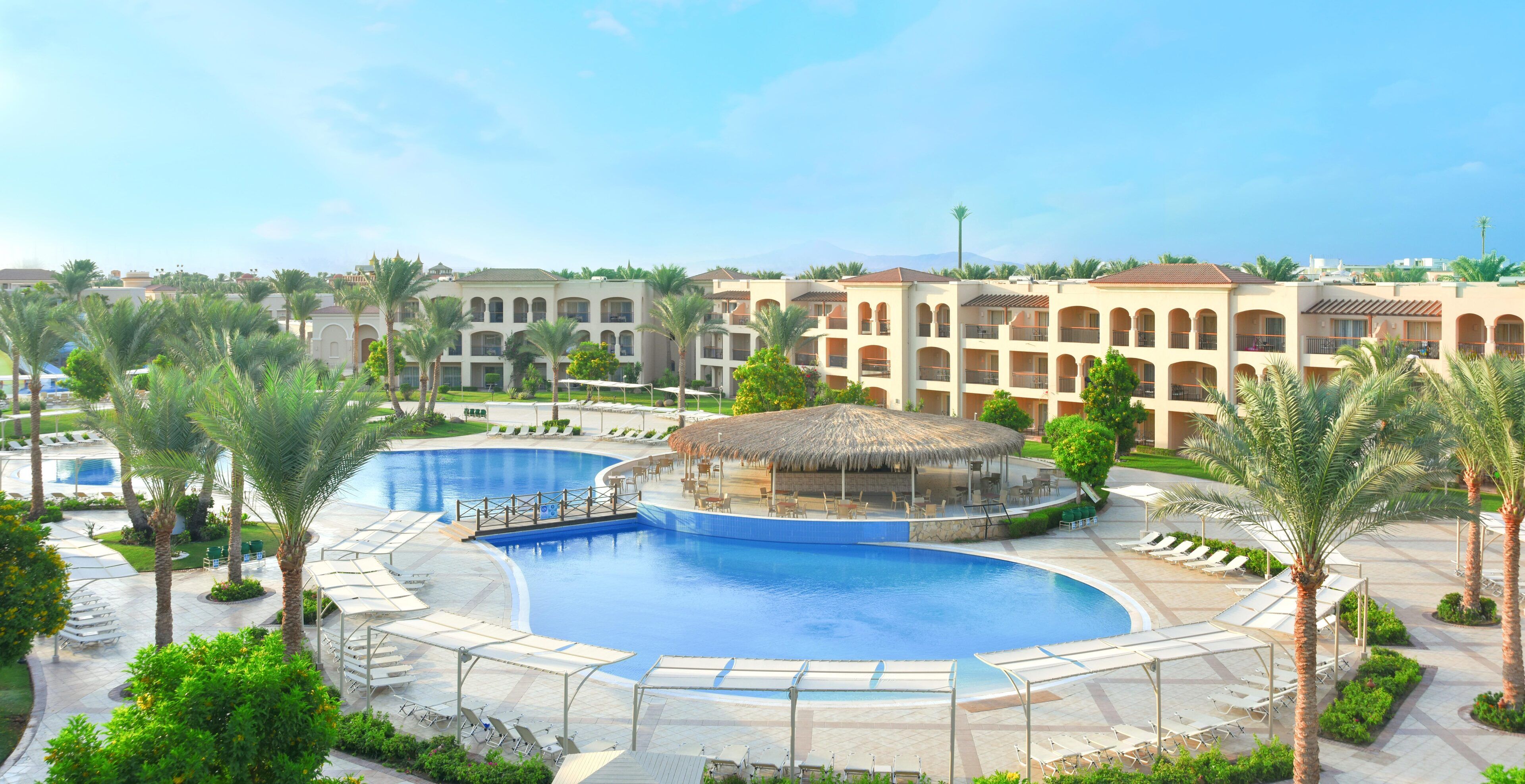 A resort with a large central pool, multiple buildings with balconies, and palm trees.