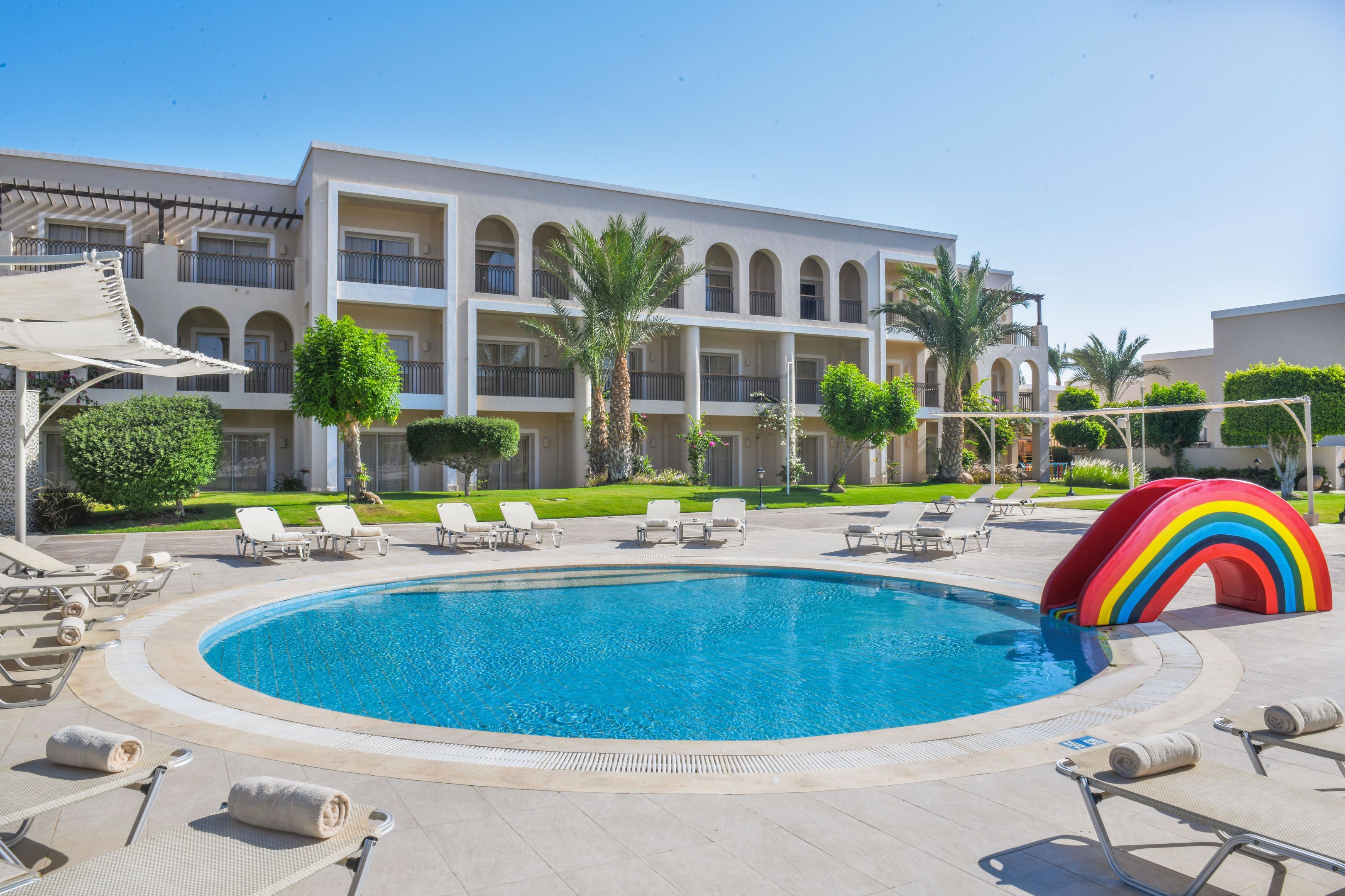 A swimming pool with lounge chairs and a rainbow decoration, surrounded by a building with balconies and palm trees.