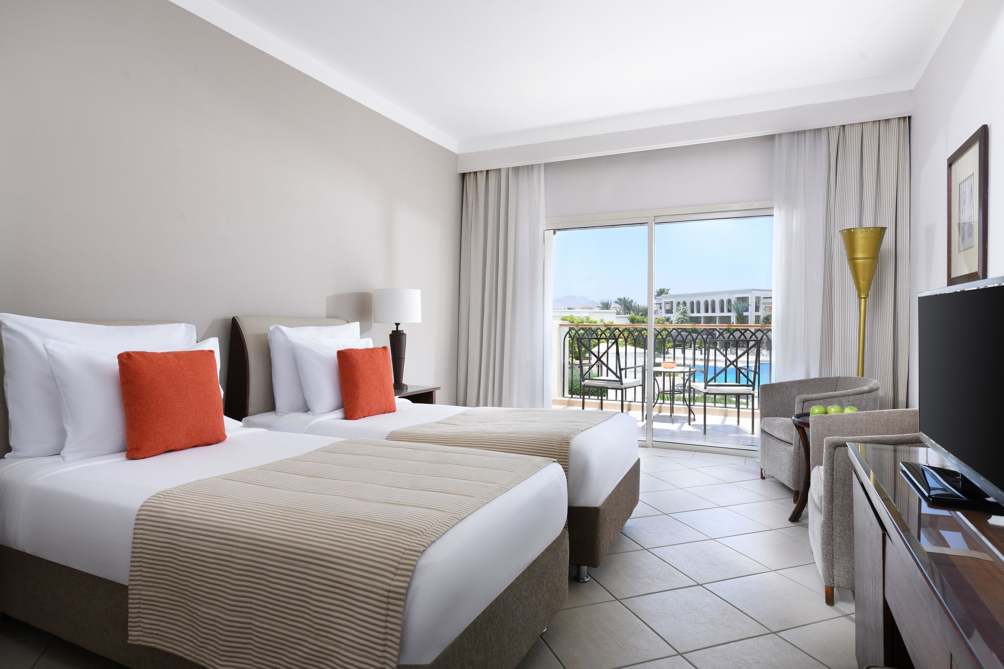 A hotel room with two beds, a TV, a balcony with a pool view, and a chair.