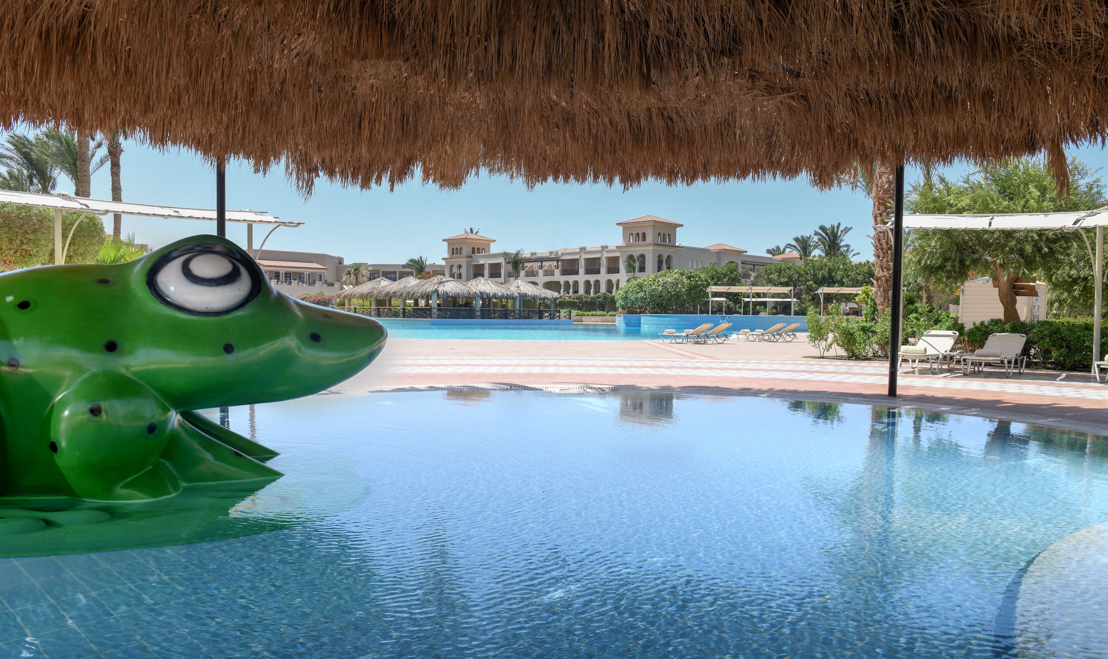 A pool with a frog-shaped water feature, lounge chairs, and a building with a thatched roof in the background.