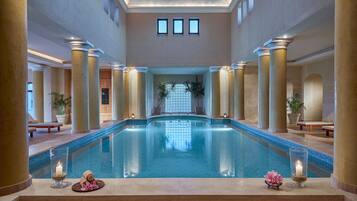 A large indoor swimming pool with a view of a wooden structure in the background, surrounded by columns and potted plants.