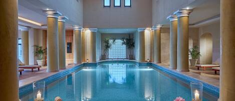 A large indoor swimming pool with a view of a wooden structure in the background, surrounded by columns and potted plants.
