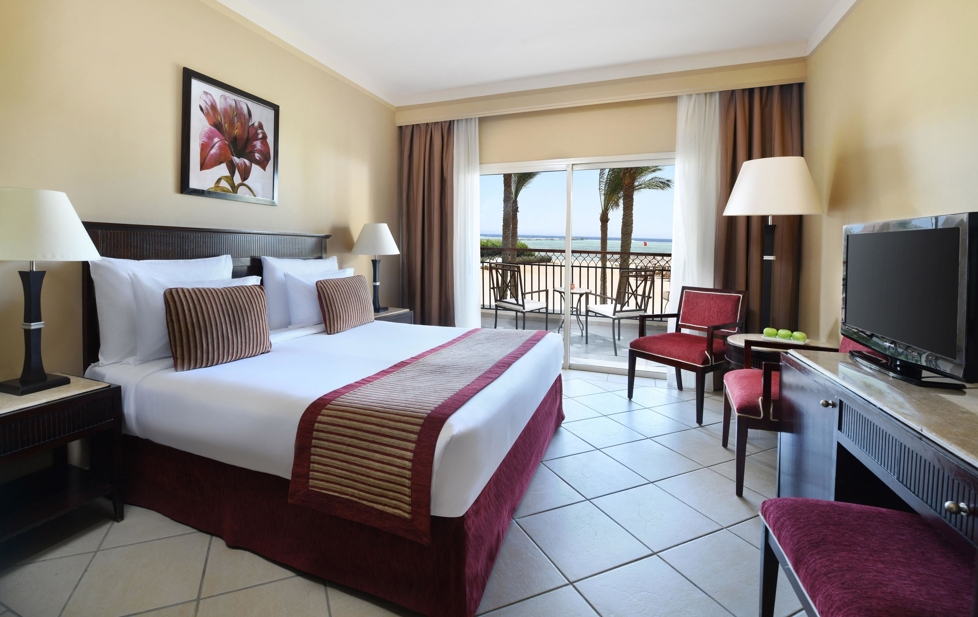A hotel room with a bed, a television, a desk with a computer, and a balcony with a view of the ocean.