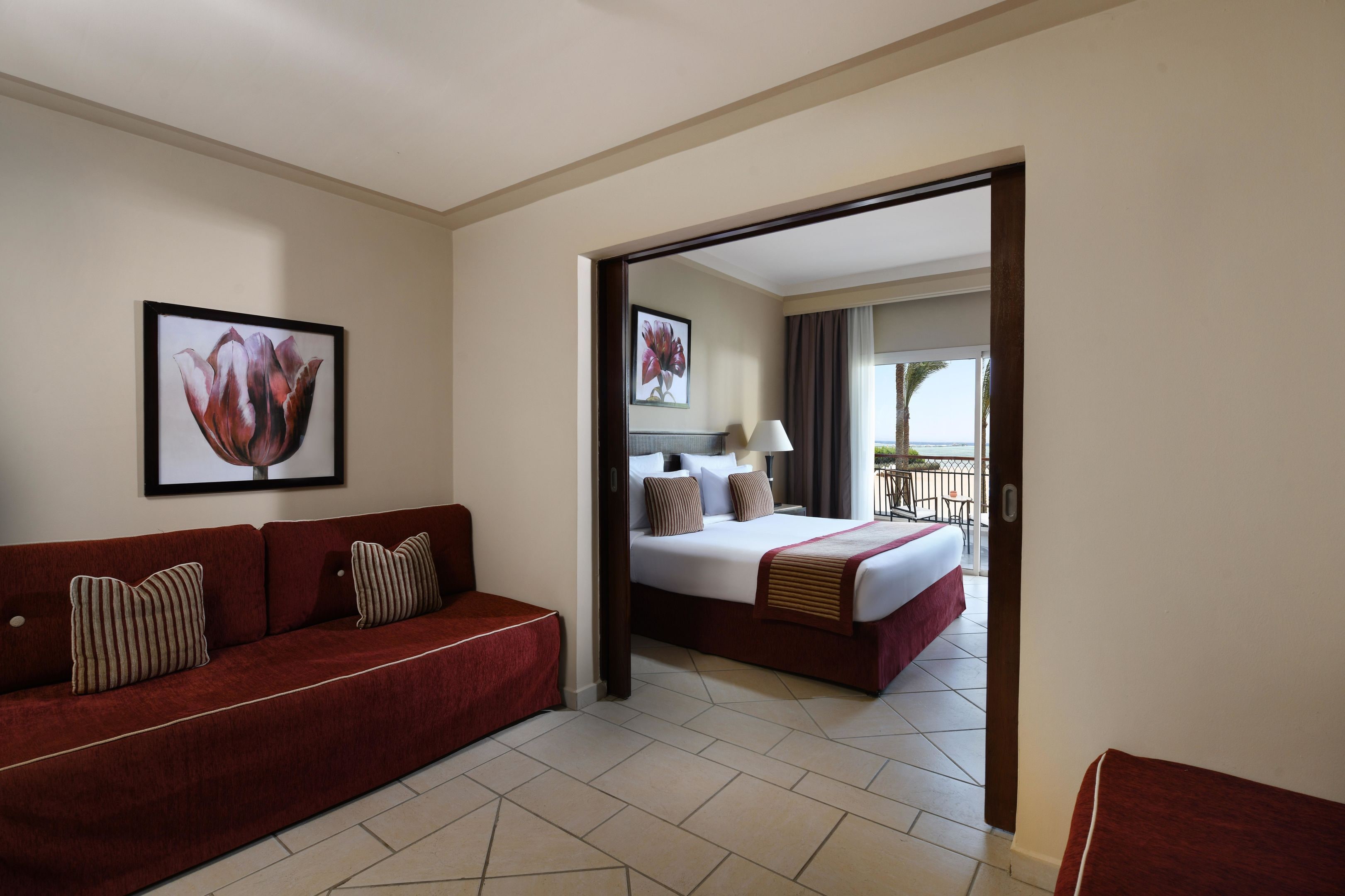 A hotel room with a red sofa, a bed with white bedding, a framed floral artwork, and a view of the balcony.