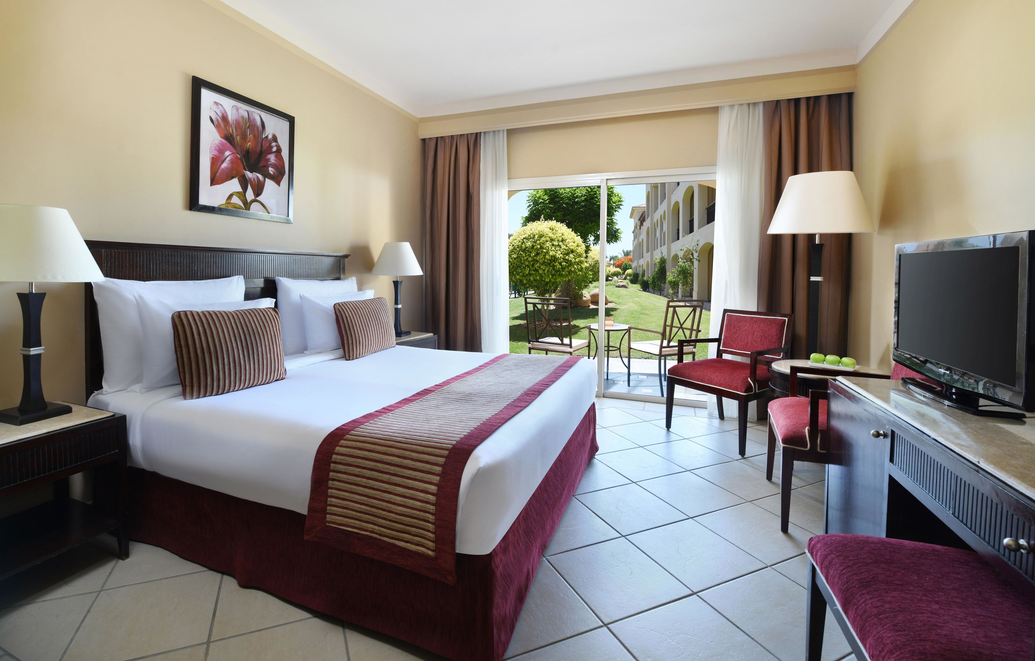 A hotel room with a large bed, two bedside tables with lamps, a television, a desk with a chair, and a view of the garden through the window.