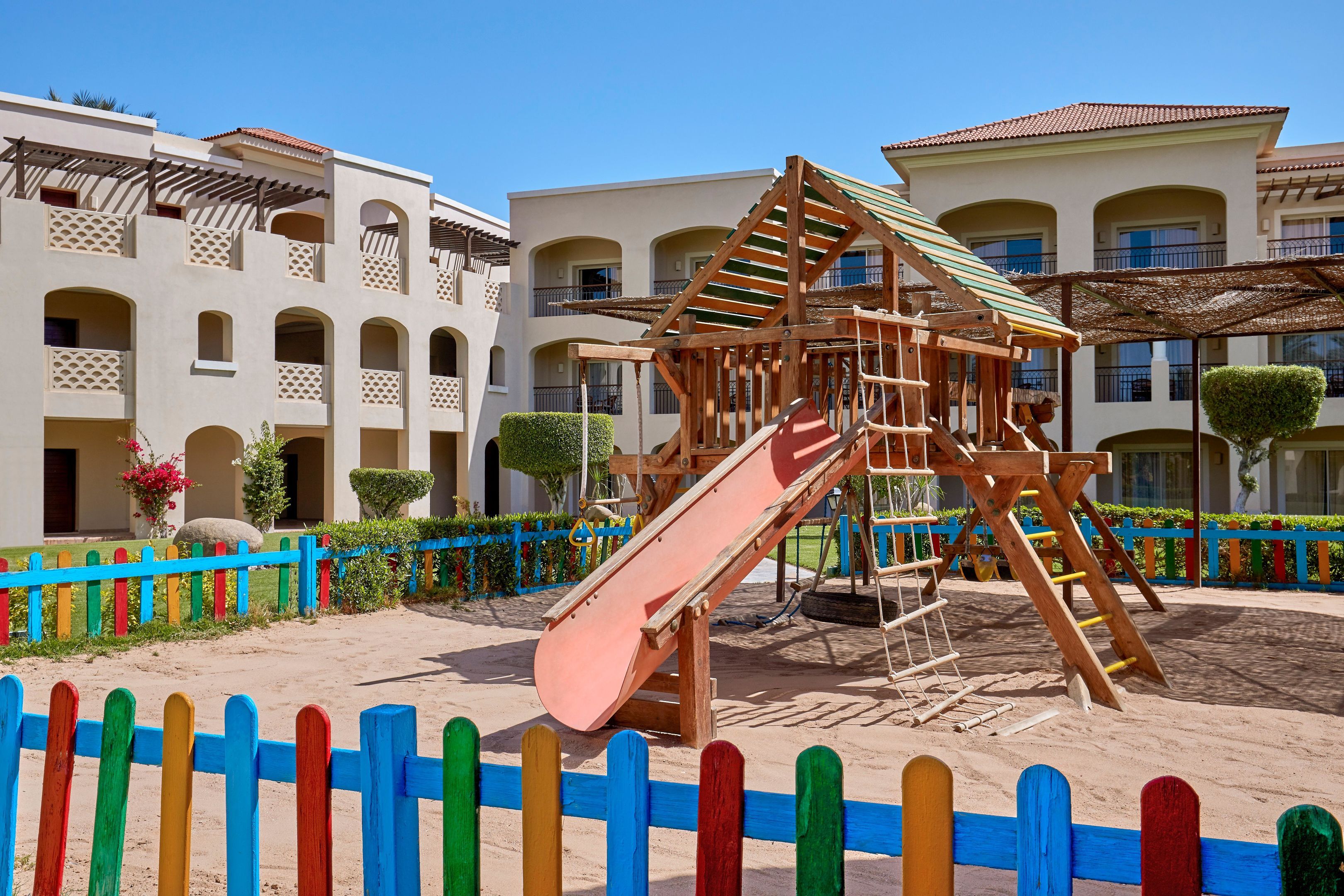 Un parque infantil con tobogán y estructura de escalada, rodeado de una valla de colores y un edificio con varios balcones.