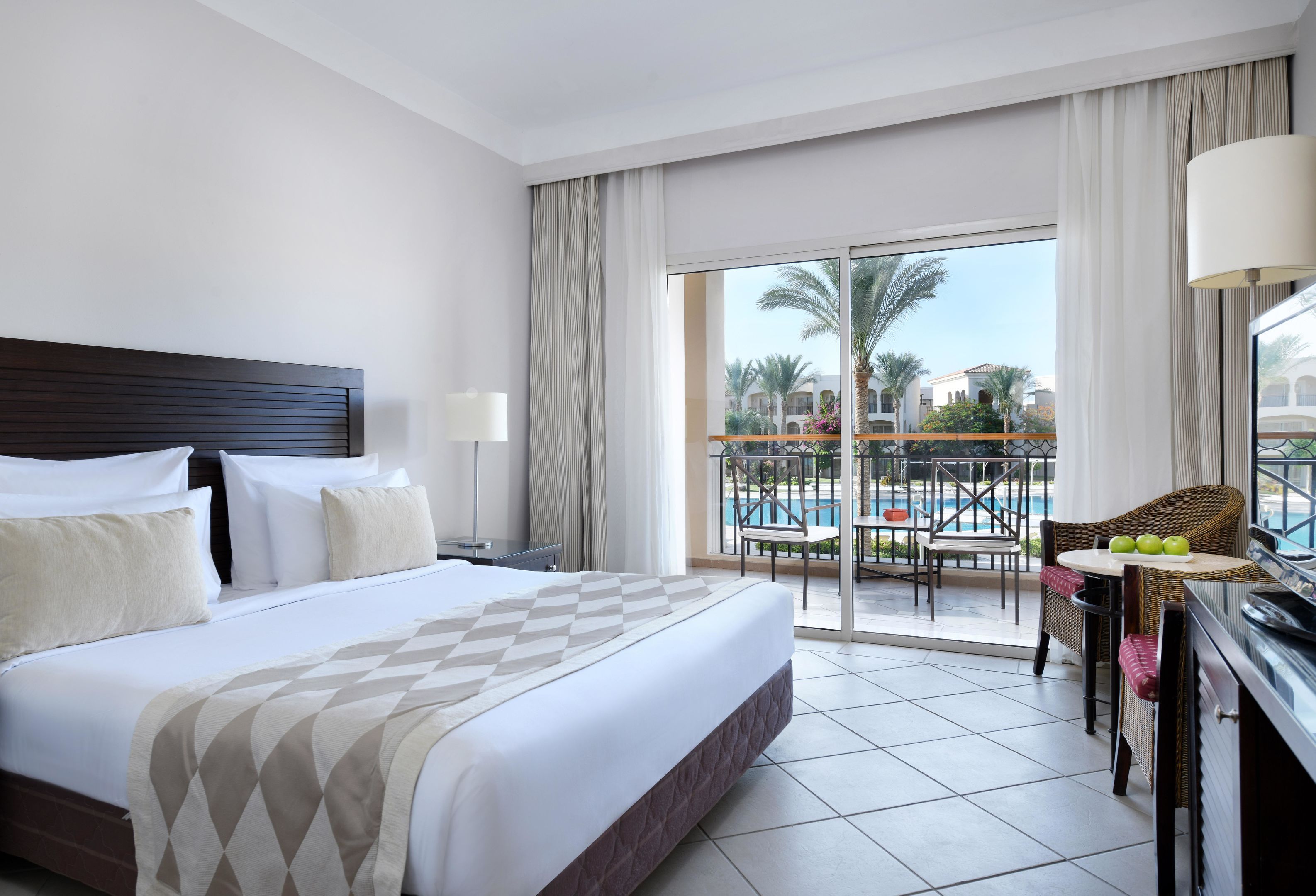 A hotel room with a large bed, a balcony with a pool view, and a dining area with chairs and a table.