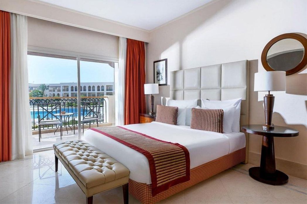 A hotel room with a large bed, a bench, a round table, a nightstand, a lamp, a mirror, and a view of a pool and buildings.