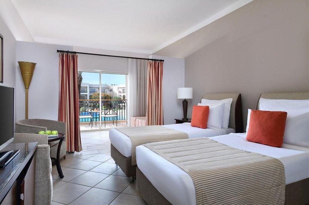 A hotel room with two beds, a desk, a chair, a lamp, and a view of the outside.