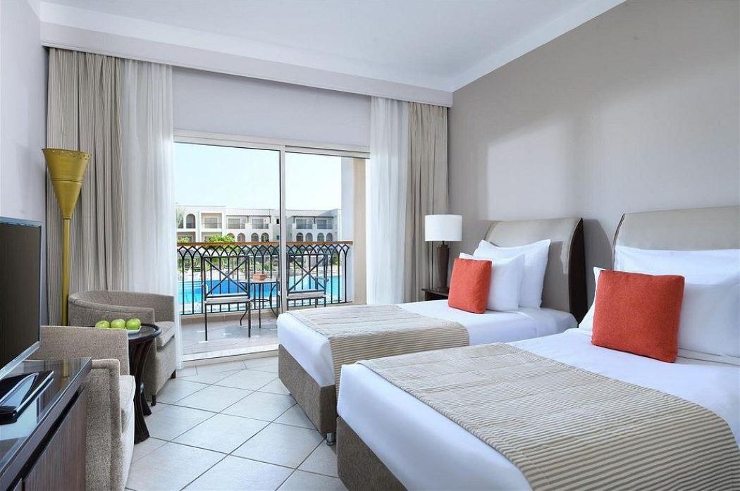 A hotel room with two beds, a TV, a small table with apples, and a balcony with a pool.