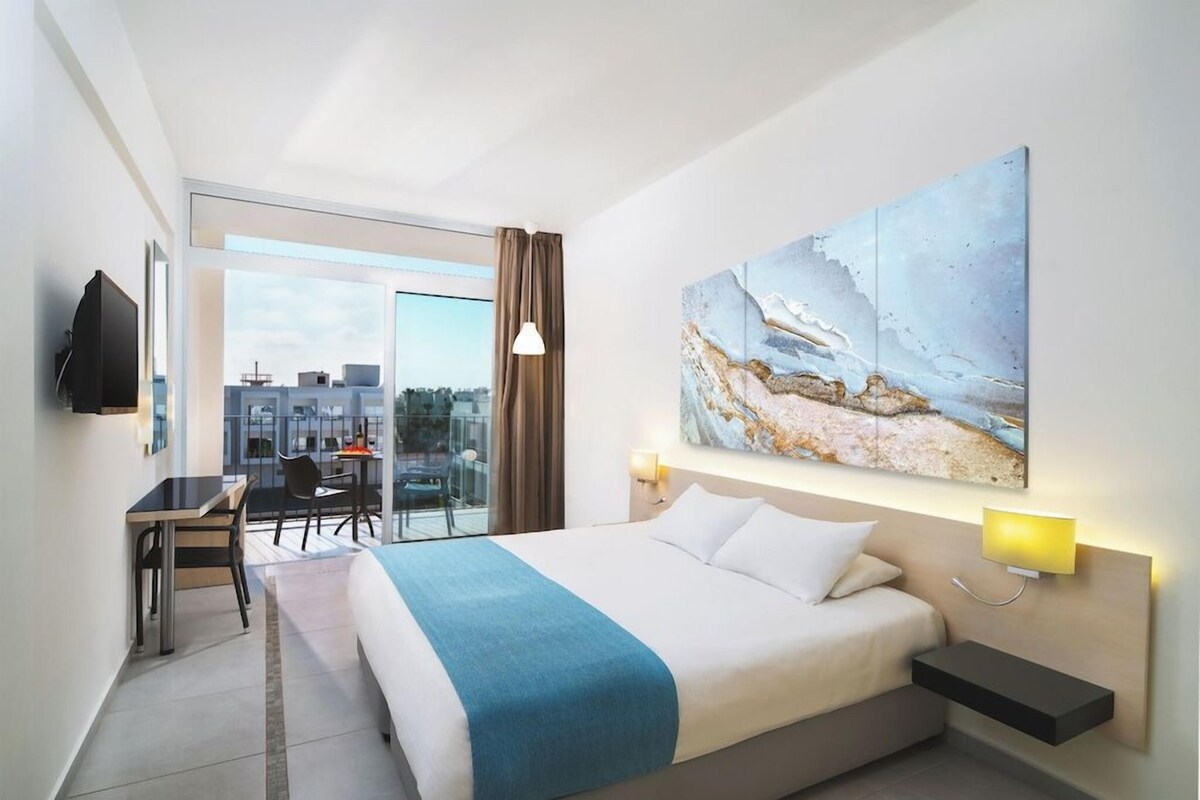 A hotel room with a bed, desk, chair, and a large window.