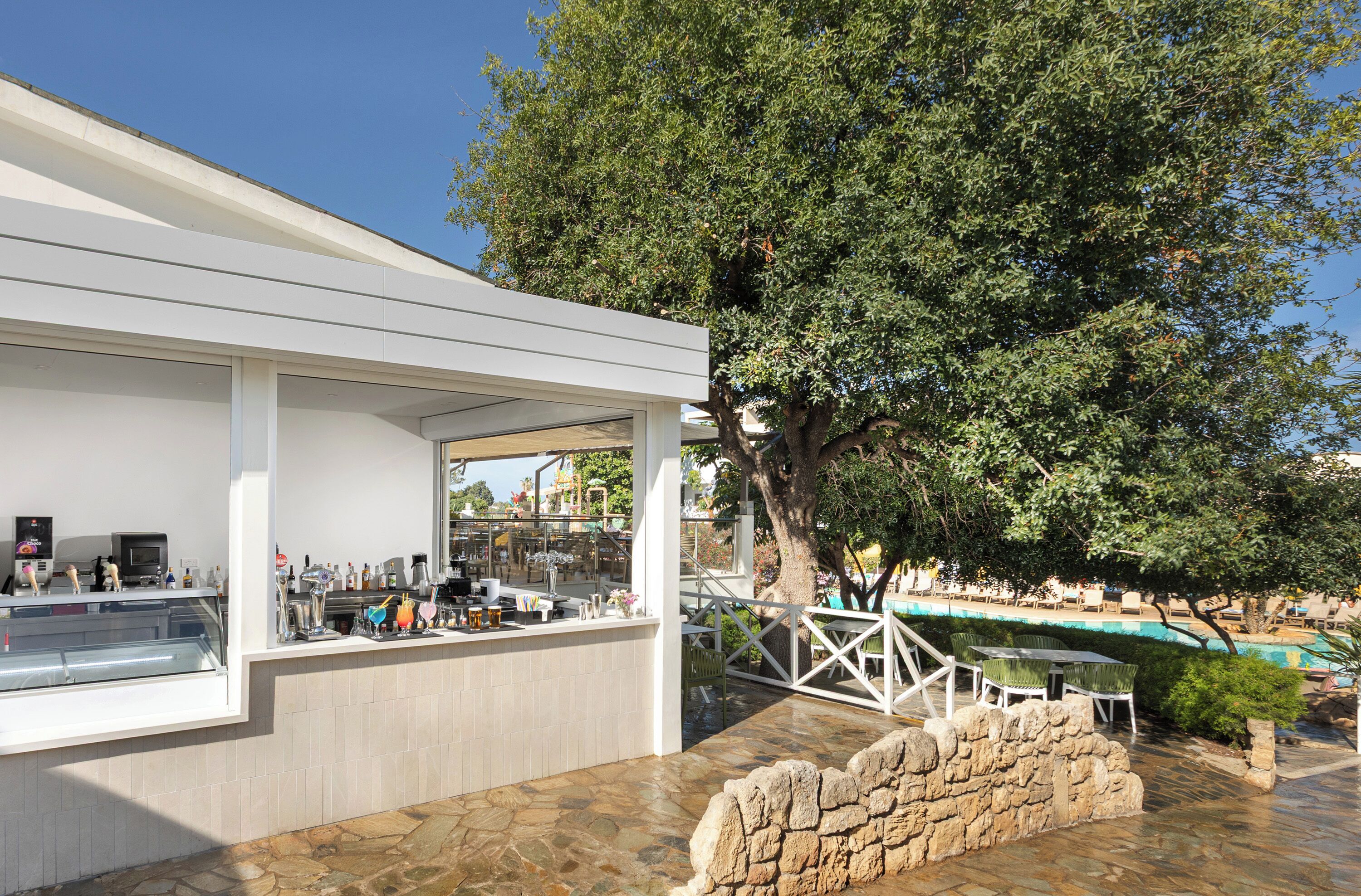 Un área de bar moderna al aire libre con una pared de piedra, un árbol grande y una piscina al fondo.