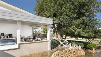Un área de bar moderna al aire libre con una pared de piedra, un árbol grande y una piscina al fondo.