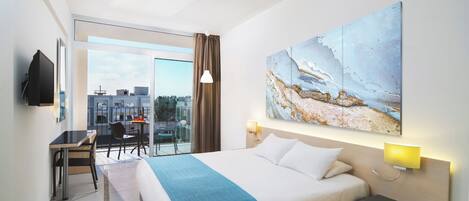 Habitación de hotel con cama, escritorio y silla; un ventanal con vistas a edificios; y un cuadro en la pared.
