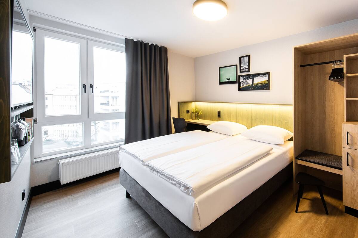 Una habitación de hotel con cama, escritorio, armario y ventana con cortinas.
