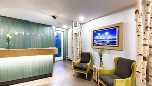 Una zona de recepción con un mostrador de madera, dos sillones de mimbre, un televisor enmarcado en la pared y cortinas con estampado de abedul.