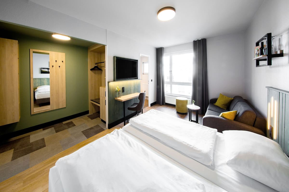 Habitación de hotel con una cama grande, un escritorio con silla, televisor, sofá con cojines y una ventana con cortinas.