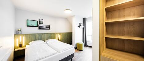 Ett hotellrum med en säng, sängbord, väggmonterade lampor, en liten växt och ett fönster med gardiner.