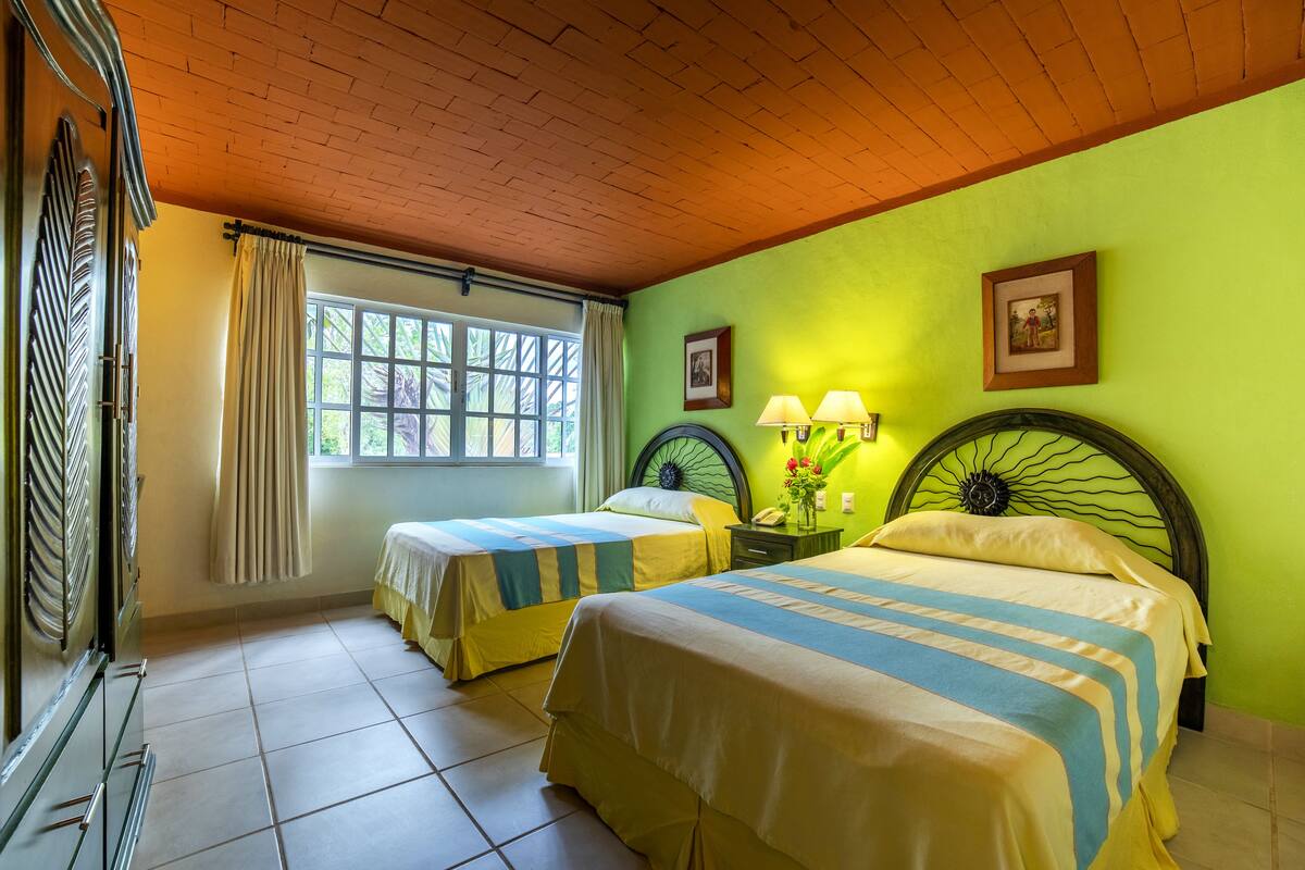 Ein Zimmer mit zwei Betten, einem Vorhangfenster, einer Holzbalkendecke und einer grünen Wand.